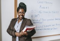 Cheerful black female teacher with workbooks standing near whiteboard